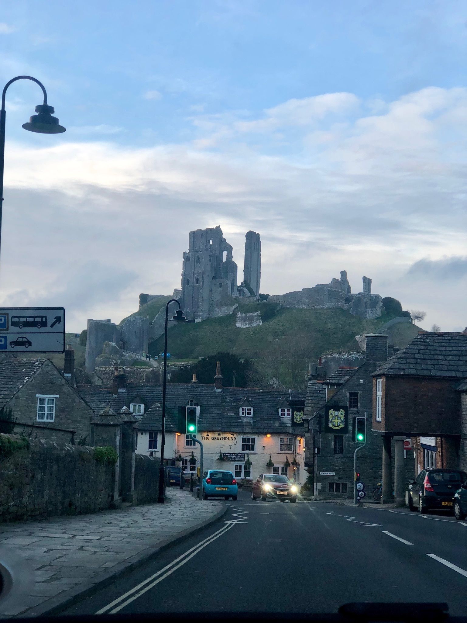 A street view with a historic castle ruin in the background, a pub named 'The Greyhound' in the foreground, and a traffic light indicating a green signal.