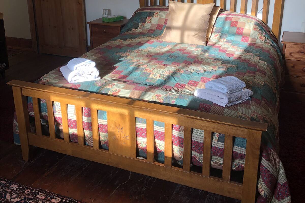 Una cama de madera con colcha, una mesita de noche con lámpara y una puerta en una habitación con alfombra estampada.