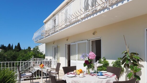 Zona pranzo all'aperto con sedie in vimini e un tavolo apparecchiato per due, con vista su un edificio bianco con balconi.