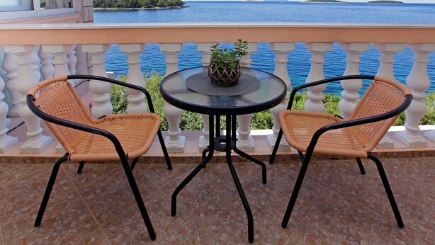 A balcony with two wicker chairs and a small round table, overlooking a calm sea and distant islands.