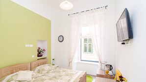 A bedroom with a bed, a bedside table, a TV mounted on the wall, a window with curtains, and a clock on the wall.