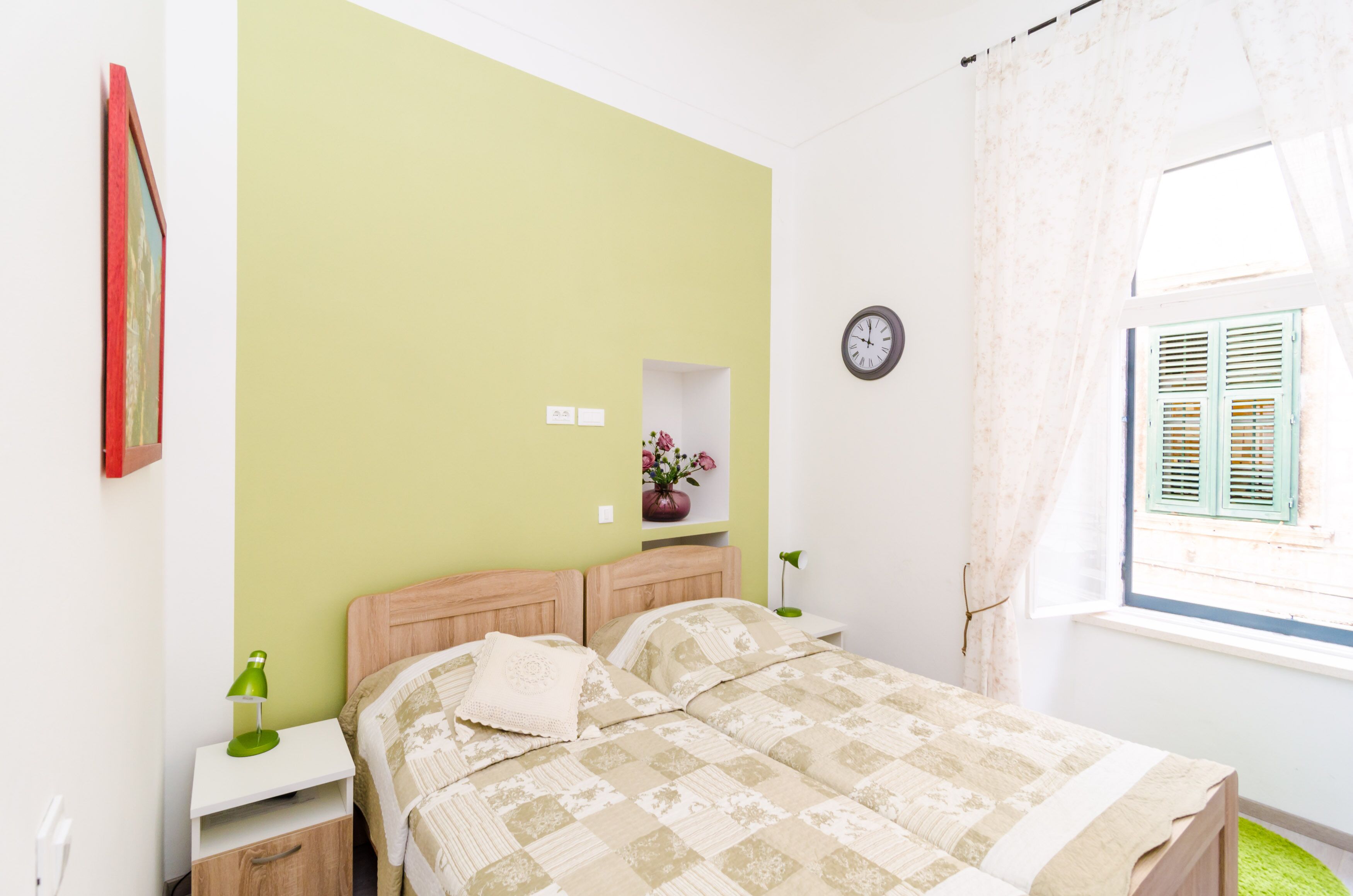 Un dormitorio con cama, mesitas de noche, un reloj y una ventana con contraventanas.
