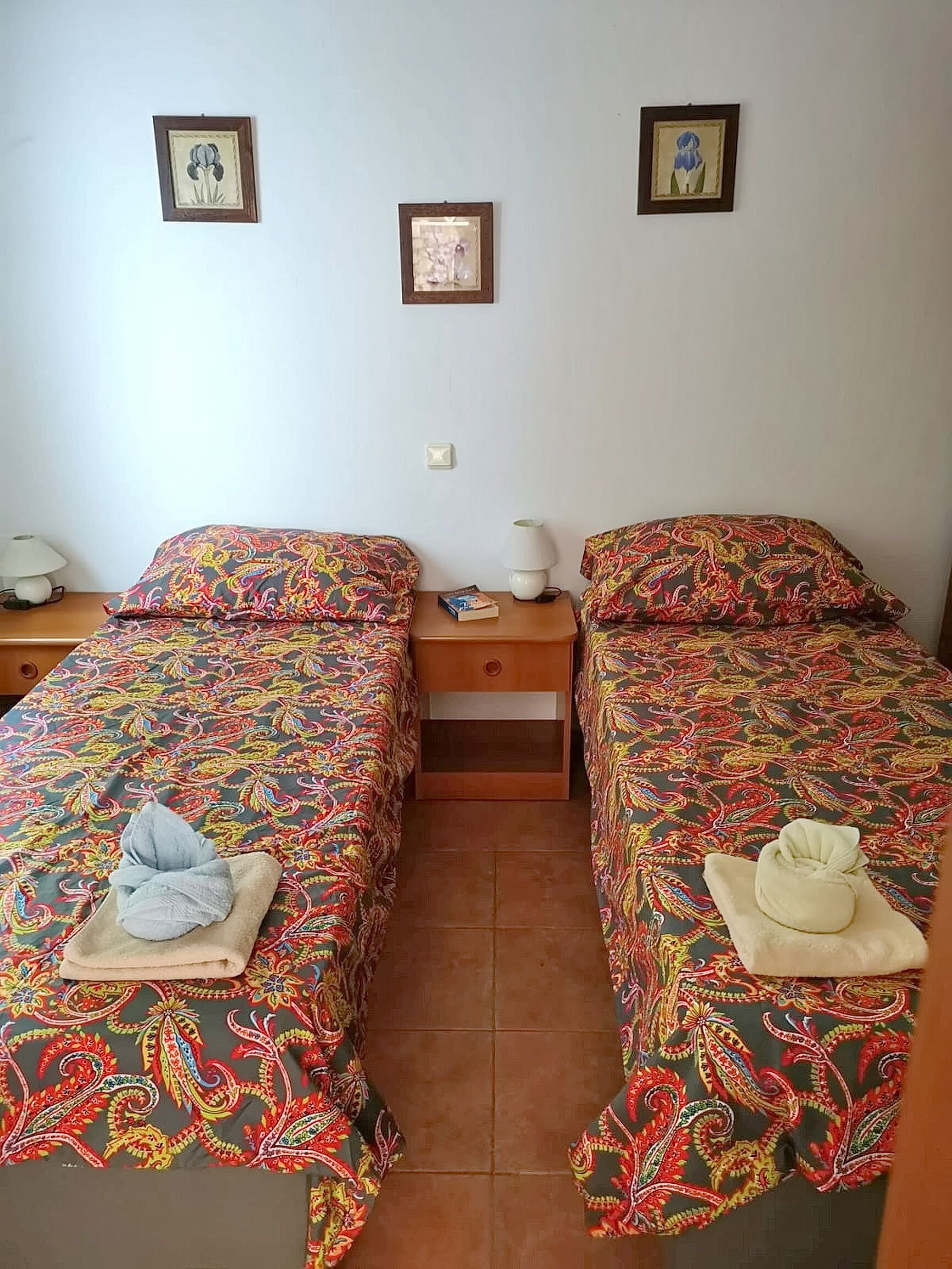 A room with two beds, each with a colorful patterned bedspread and a towel on the bed. There are two nightstands with lamps and a small table between the beds.