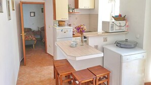Cocina compacta con refrigerador, microondas y un lavamanos pequeño. Hay una mesa de madera y una silla en el centro, y una ventana con una cesta sobre la encimera.