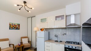 Une cuisine compacte équipée d’une cuisinière à gaz, d’un four et de meubles blancs. On y trouve un petit coin repas avec deux chaises et une table, ainsi qu’un tableau accroché au mur.