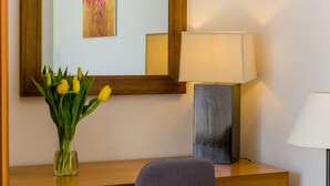 Une chambre d’hôtel avec un bureau en bois, un vase rempli de tulipes jaunes, une lampe moderne et un grand miroir qui reflète un tableau aux couleurs vives.