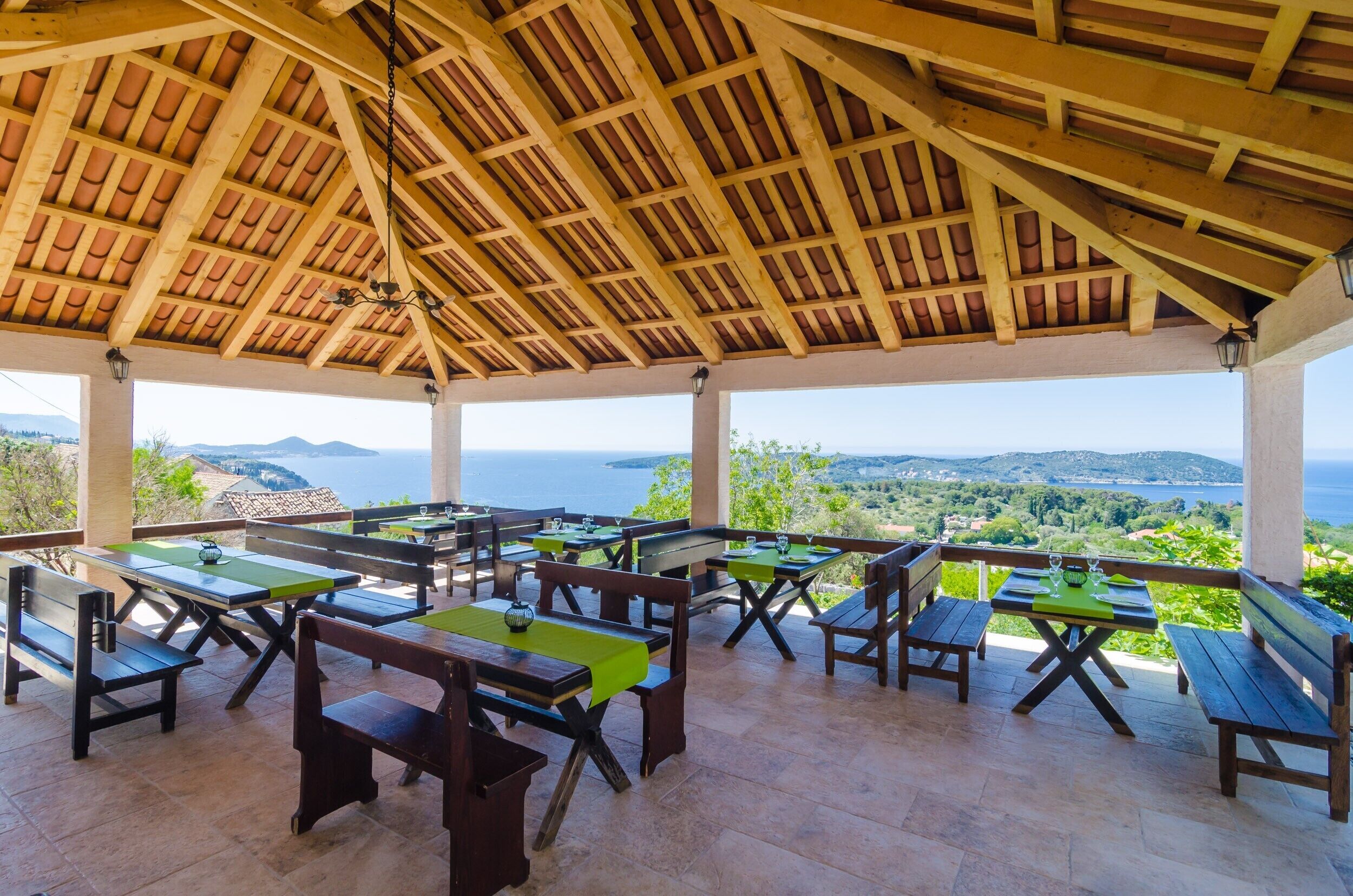 An outdoor dining area with wooden tables and benches, a tiled floor, and a view of the sea and landscape.
