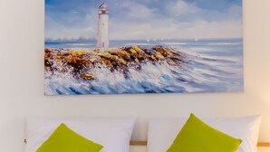 Quarto com cama, almofadas brancas e verdes, pintura de um farol na parede e tomada visível.