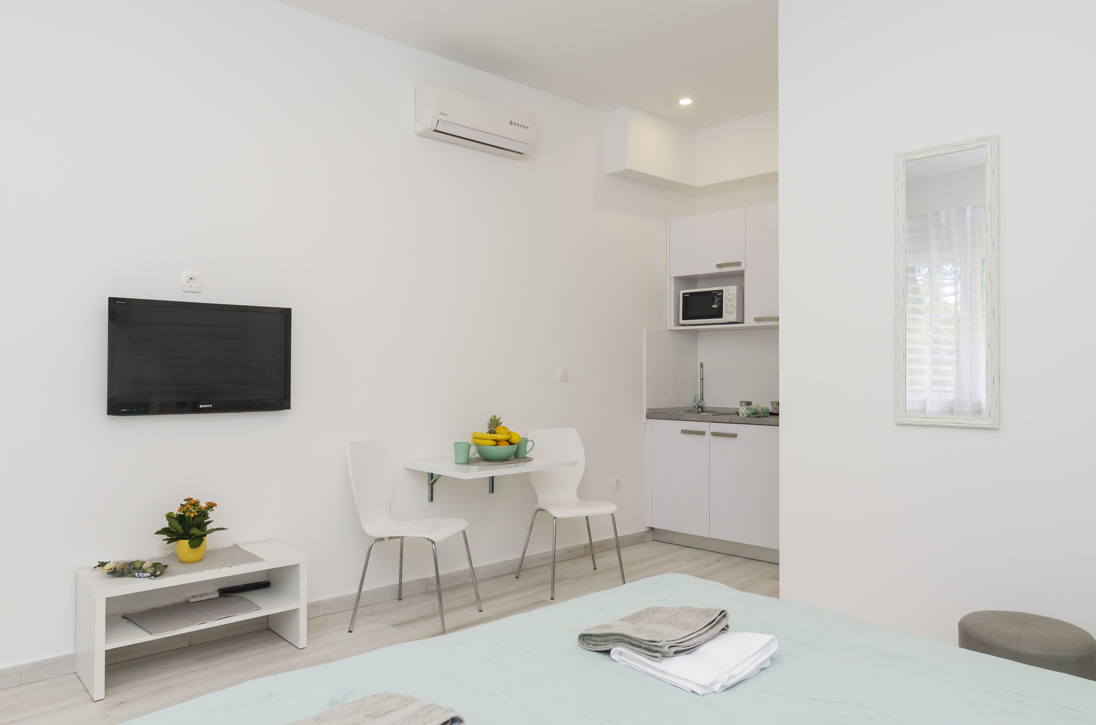 Una habitación moderna y minimalista con televisión de pantalla plana en la pared, una pequeña mesa de comedor con sillas, una kitchenette con microondas y un amplio ventanal con cortinas.