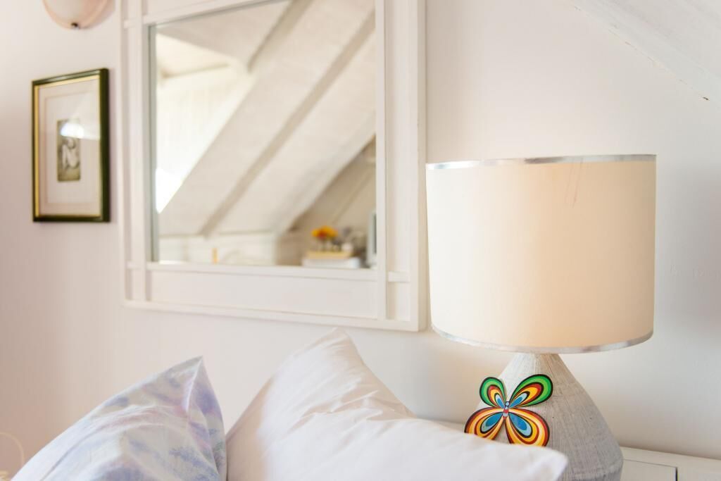 A bedroom with a bed, a bedside lamp with a colorful base, a framed picture on the wall, and a window with a view of the interior.