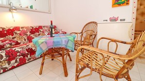 Ein gemütlicher Wohnbereich mit einem blumengedeckten Sofa, einem Tisch mit bunten Tischdecken und Korbstühlen, sowie einem Kühlschrank mit Dekoration.