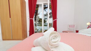 Ein Hotelzimmer mit einem Bett, einem Holzschrank, einem roten Vorhang, einem weißen Stuhl und Blick auf Bäume durch eine Glastür.