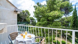 A balcony with a table set for two, overlooking a garden with pine trees and a clear sky.