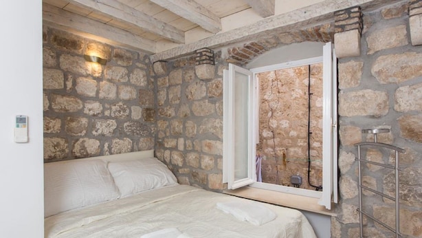 Une chambre avec un mur en pierre, un lit recouvert de draps blancs, une fenêtre donnant sur un beau paysage et un porte-serviettes en métal.