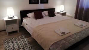 A neatly made bed with a beige comforter, two pillows, and a folded towel on the side. There are two bedside tables with lamps, a patterned rug, and two framed pictures on the wall above the bed.
