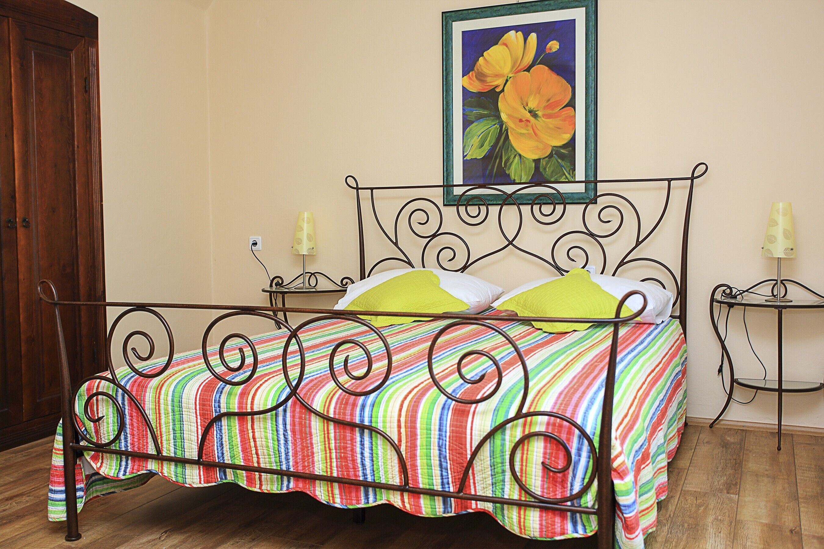 Een bed met metalen frame, een kleurrijke streepdeken en twee kussens. Boven het bed hangt een schilderij van bloemen aan de muur.