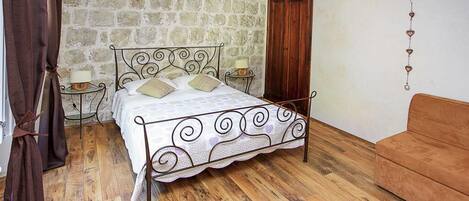 Een slaapkamer met een stenen muur, een bed met een decoratieve hoofdbord, een houten vloer, een houten deur, een klein nachtkastje met een lamp en een bruine bank.