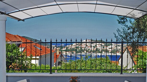 An outdoor patio with wicker furniture, a glass table, and a view of a coastal town through a metal fence.
