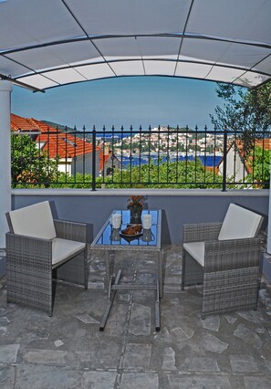 Um pátio externo com móveis de vime, uma mesa de vidro e vista para uma cidade costeira através de uma grade de metal.