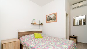 Une chambre à coucher comprenant un lit, une table de chevet, une petite armoire en bois, une étagère avec deux lampes, un tableau encadré au mur et un miroir.