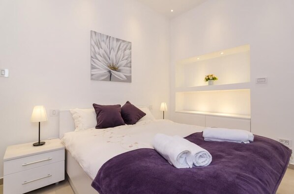 Ein modernes Schlafzimmer mit einem großen Bett, zwei Nachttischen, einem Wandregal mit einer Vase und einem floralen Kunstwerk.
