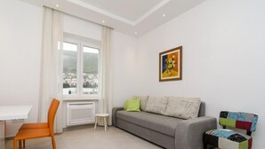 Ein modernes Wohnzimmer mit einem grauen Sofa, einem bunten Sessel, einem kleinen weißen Tisch und einem Fenster mit Blick auf Gebäude.