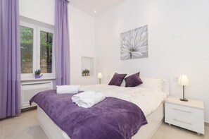 Ein Schlafzimmer mit einem Bett, weißen Nachttischen, einem Fenster mit violetten Vorhängen, einem Blumenbild an der Wand und einer Heizung.