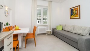 Ein modernes Wohnzimmer mit grauem Sofa, orangefarbenen Sesseln, einem weißen Esstisch und einem Fenster mit Vorhängen.