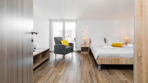 A modern bedroom with a wooden floor, a bed with white bedding and a yellow pillow, a grey armchair with a yellow pillow, a nightstand with a lamp, and a window with a view of buildings.