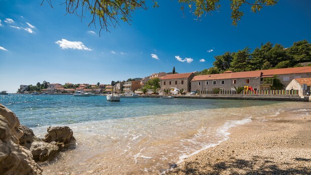 A coastal town with clear waters, a pebbly beach, and stone buildings with red roofs.