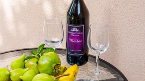 A bottle of Merlot wine, two empty wine glasses, and a bowl of green figs and a banana on a glass table against a textured wall.