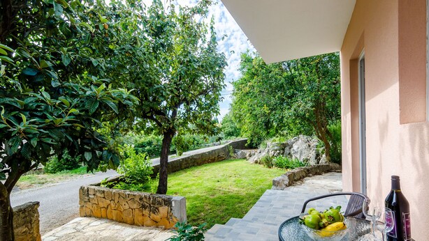 A patio with a stone wall, a glass table with a bowl of fruit, and a wine bottle.