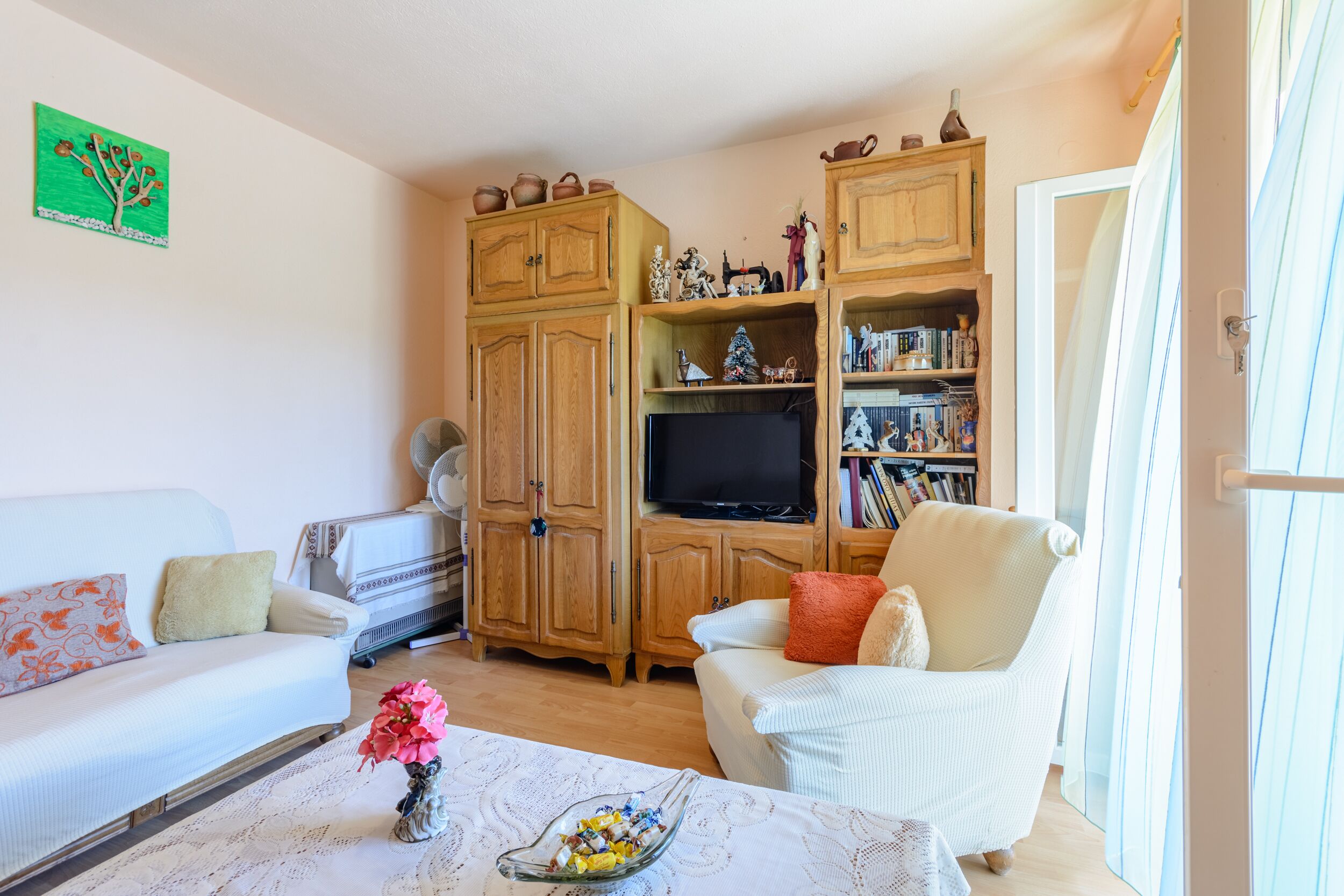Ein Wohnzimmer mit einem weißen Sofa, einem weißen Sessel, einem Glastisch mit einer Vase, Holzschränken, einem Fernseher und einem Bücherregal.