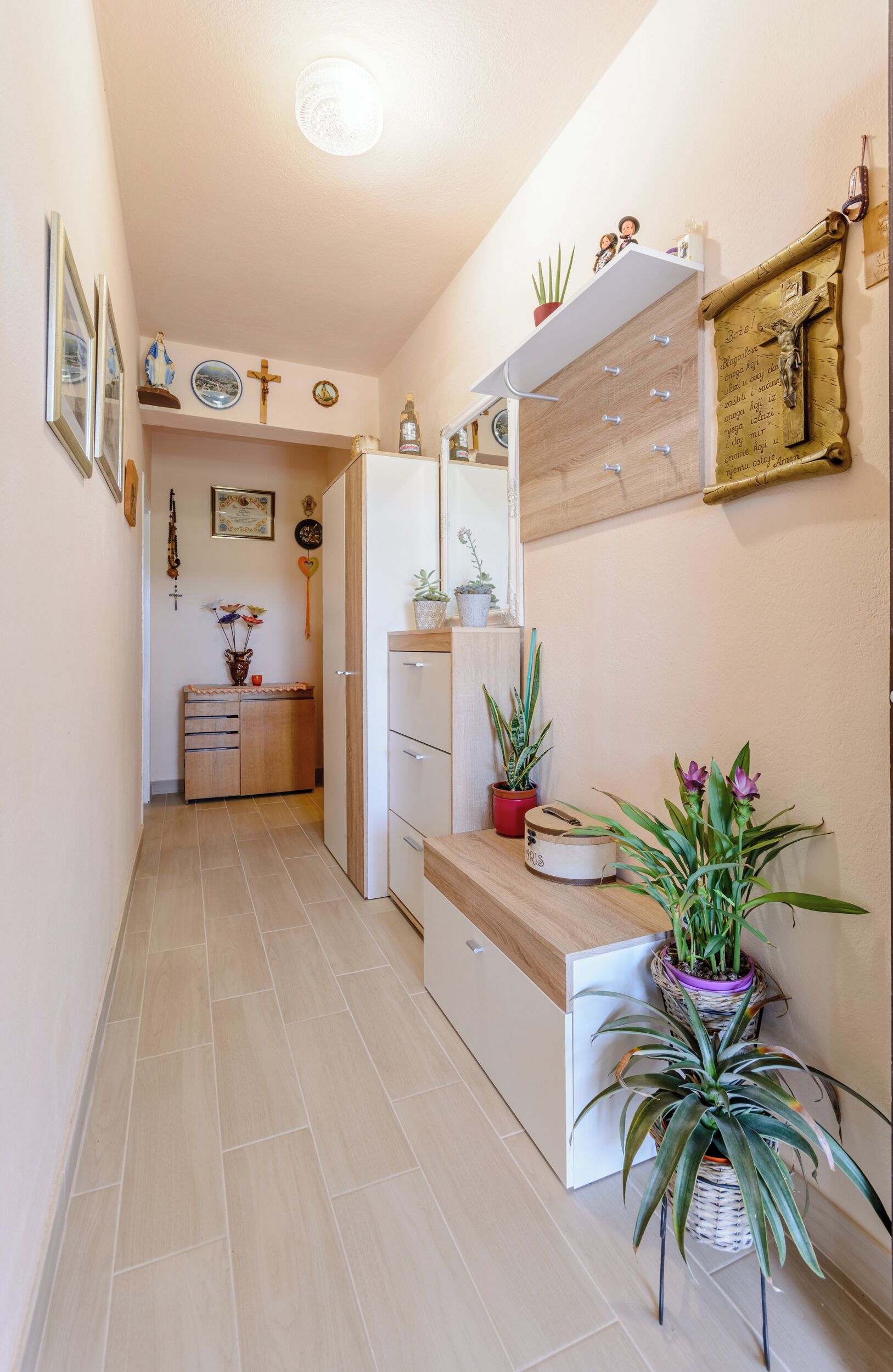 A hallway with a wooden cabinet, a shelf with plants, a mirror, and a wall with a cross and decorations.