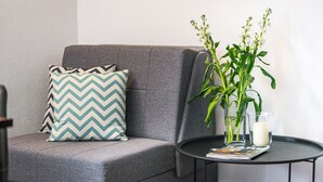 Un sofá gris con un cojín estampado, una mesita redonda con una vela y una planta, y una revista.
