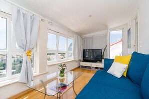 Un salón moderno con un sofá azul, una mesa de centro de cristal, un televisor de pantalla plana y grandes ventanales con vistas a la ciudad.