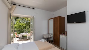 A hotel room with a bed, a TV, a wardrobe, a small table with a tablecloth, and a view of an outdoor area with a table and chairs. - Begović Guest House - Twin Room (Judy) (Dubrovnik)