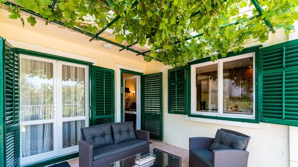 An outdoor seating area with wicker furniture and a glass coffee table, surrounded by green shutters and a pergola. - Apartment Valonija - Two Bedroom Apartment with Terrace and Garden View (Dubrovnik)