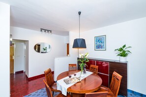 A dining area with a round wooden table, chairs, a white tablecloth, a hanging pendant light, a potted plant, and a framed picture on the wall.
