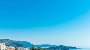 A coastal town with buildings along the shoreline, a clear blue sky, and mountains in the background. - Guest House Nikolina - Double Room with Balcony and Sea View(1) (Dubrovnik)