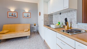 Een compacte keuken met witte kastjes, een houten aanrecht en een kleine eettafel. In de aangrenzende woonkamer staat een bank met kussens.