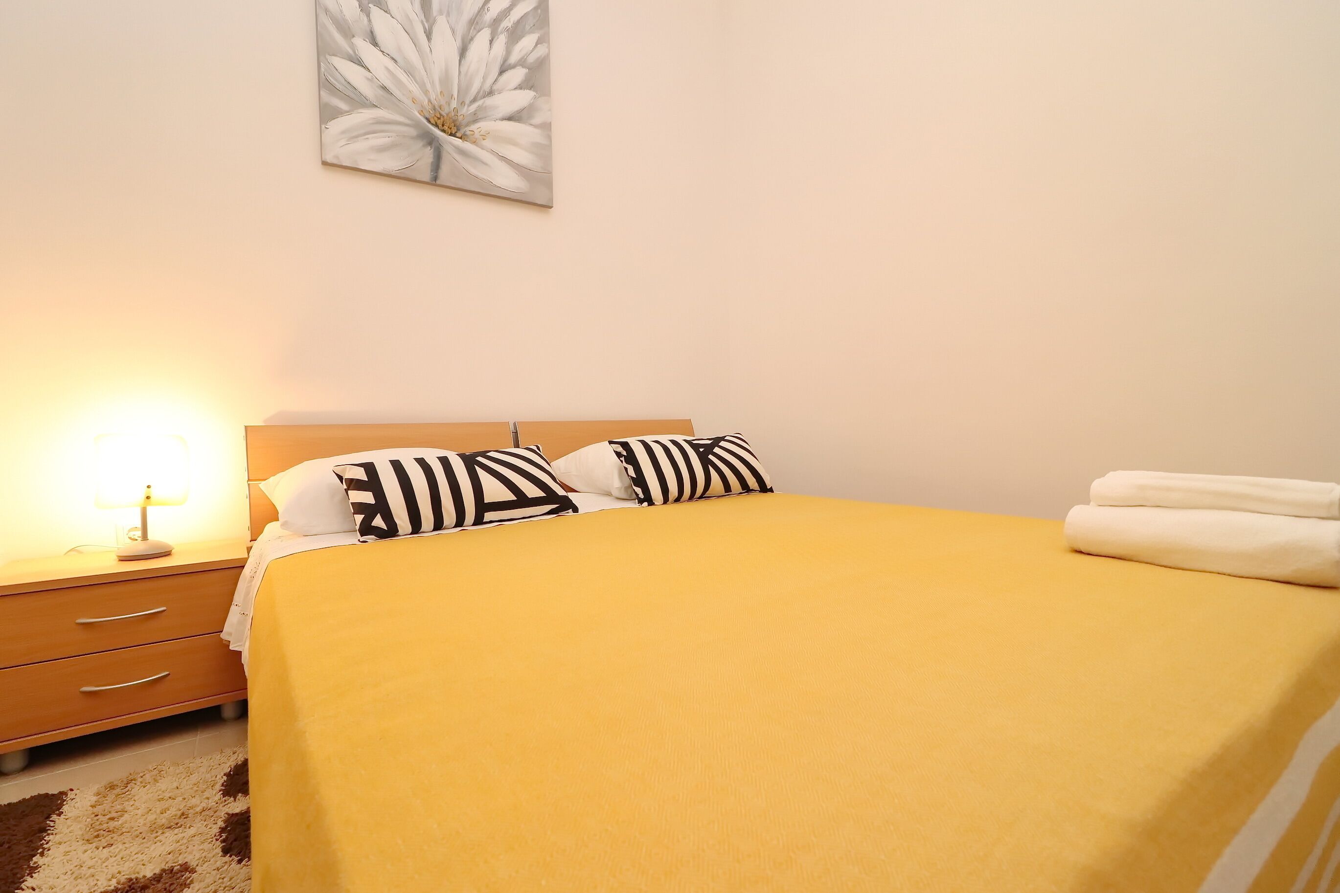 A neatly made bed with a yellow bedspread and black and white striped pillows, a bedside lamp, and a wall-mounted flower painting.