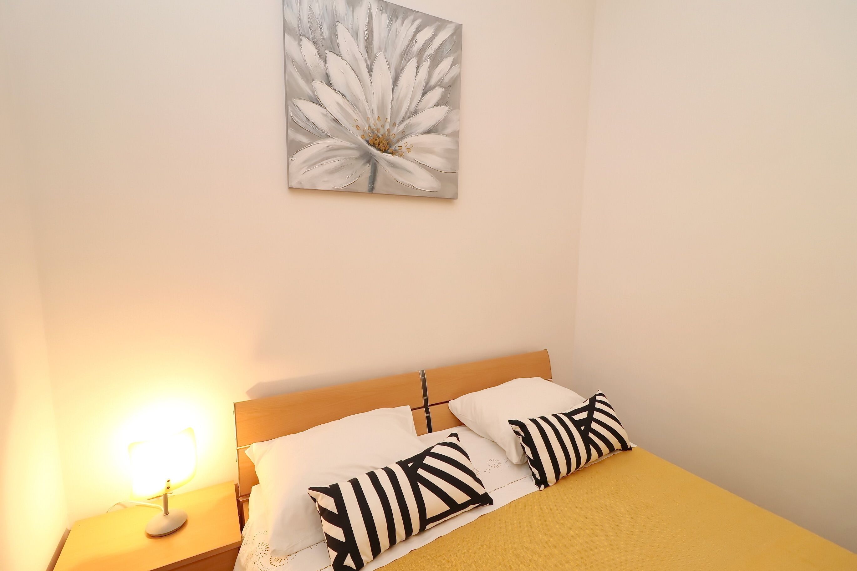 A neatly made bed with a yellow bedspread and two black and white striped pillows. A bedside lamp is on, and a painting of a flower hangs above the bed.