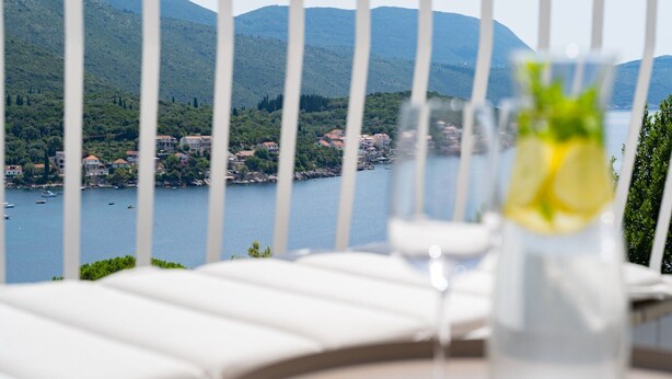 A balcony with a clear view of a lake and mountains, a glass of water with a lemon slice, and white lounge chairs.