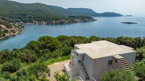 A coastal building with a flat roof, surrounded by lush greenery and a view of a calm sea with boats.