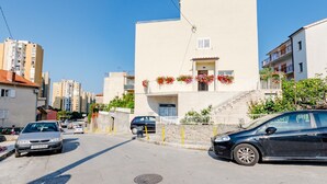 A street view with a beige building featuring a balcony with red flowers, a staircase, and a parked black car. - Apartment Marko (ST) - Three Bedroom Apartment with Balcony and Garden View (Split)