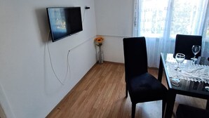 A room with a flat-screen TV mounted on the wall, a small table with two chairs, a vase with a sunflower, and a window with sheer curtains. - Apartment Marko (ST) - Three Bedroom Apartment with Balcony and Garden View (Split)