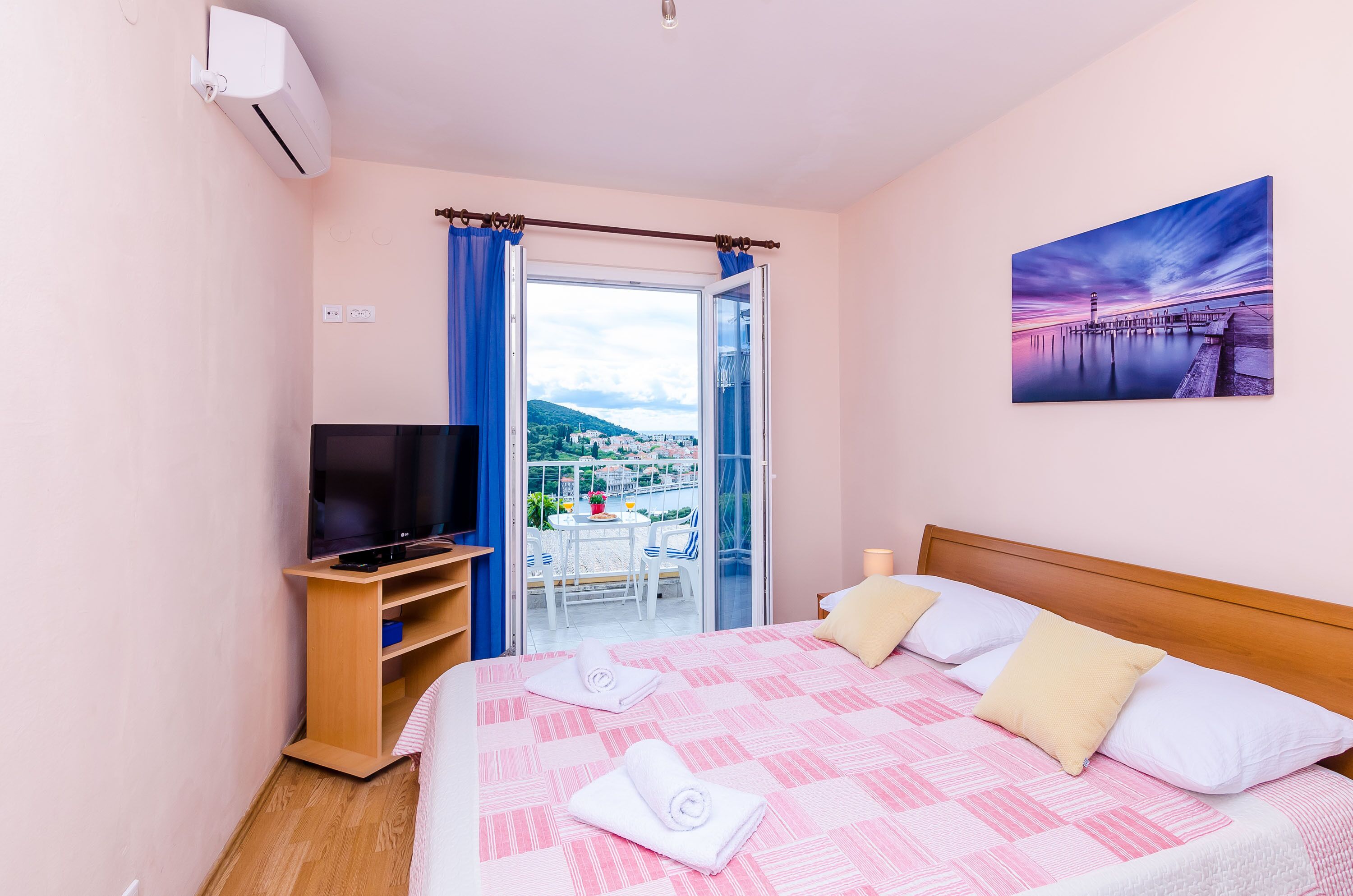 Habitación de hotel con cama, televisión, balcón con mesa y sillas, y un cuadro en la pared.