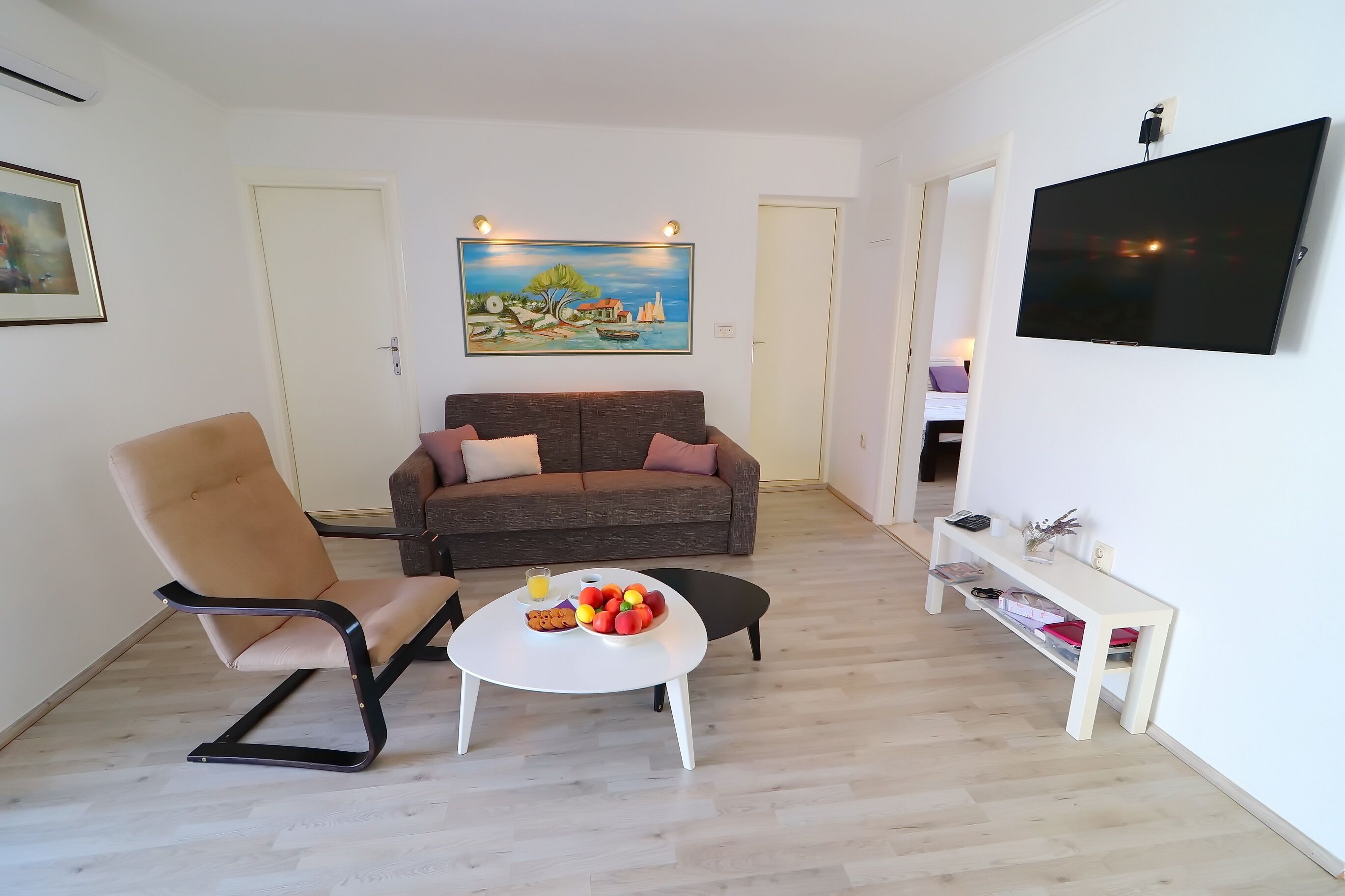 A living room with a sofa, a chair, a coffee table with fruit, and a TV on the wall.
