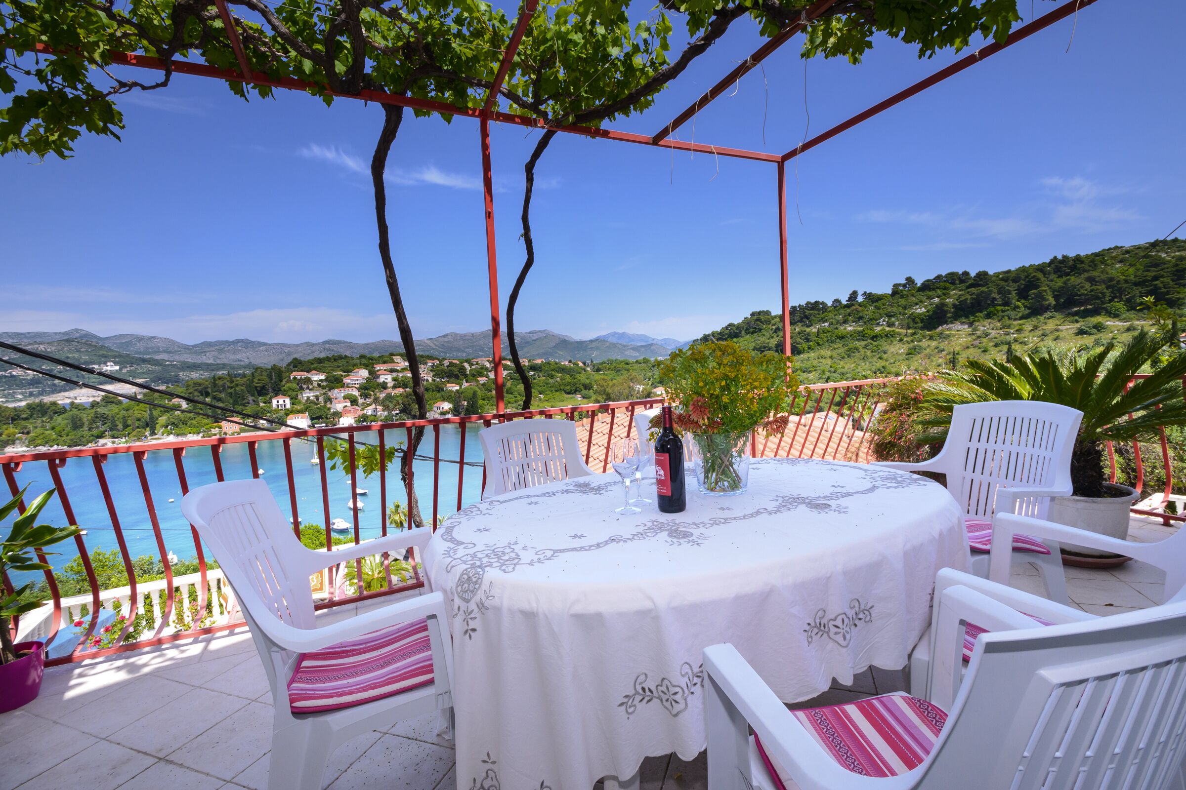 A round table with a white tablecloth and chairs with pink and white cushions, set on a balcony overlooking a body of water and a mountainous landscape.